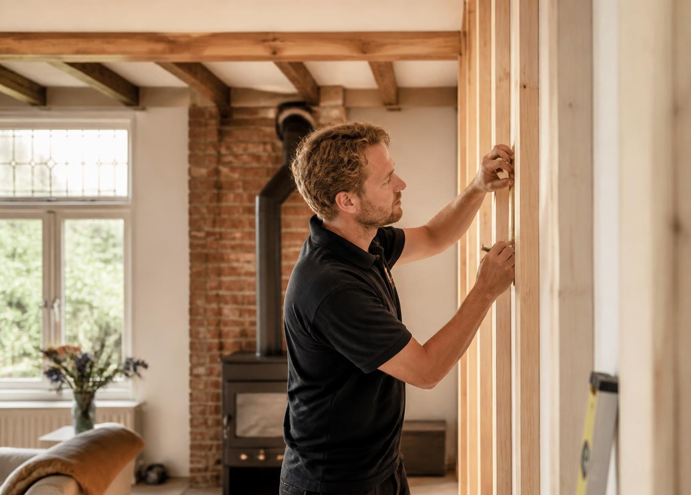 Vakman in donkere polo werkt met aandacht aan een houten wandframe in een lichte woonkamer met oude balken en baksteen